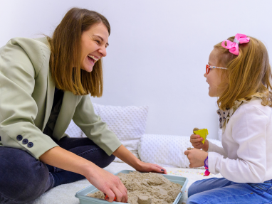 An adult woman and a young girl sit on the floor, smiling and playing with kinetic sand in a tray. The girl wears glasses and a pink bow, and both appear happy and engaged in the activity.
