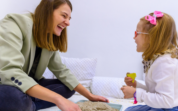 An adult woman and a young girl sit on the floor, smiling and playing with kinetic sand in a tray. The girl wears glasses and a pink bow, and both appear happy and engaged in the activity.