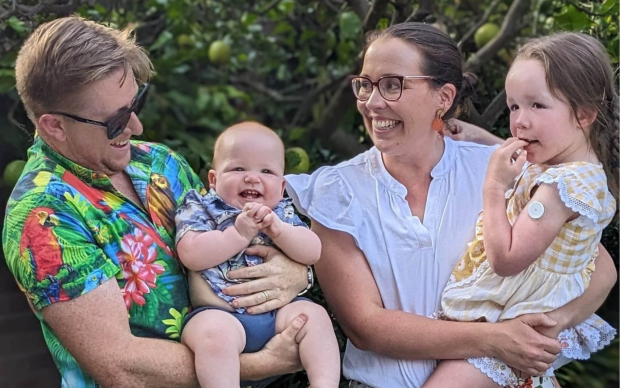 A smiling family of four stands outdoors, surrounded by greenery. The father holds a laughing baby, and the mother holds a young girl with a medical patch on her arm. All are happy and casually dressed.