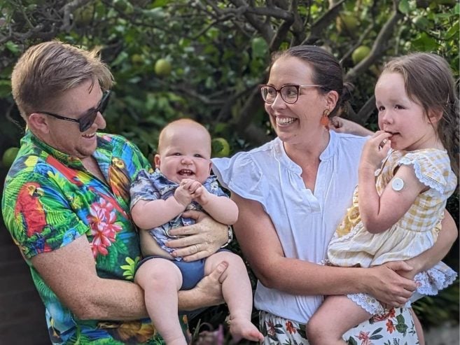 A smiling family of four stands outdoors, surrounded by greenery. The father holds a laughing baby, and the mother holds a young girl with a medical patch on her arm. All are happy and casually dressed.