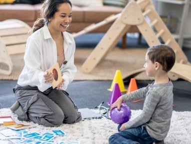 A woman sits on the floor holding a flashcard, smiling at a young boy who is holding a purple sensory ball. They are in a playroom with toys, cones, and activity cards around them.