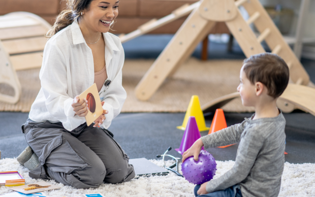 A woman sits on the floor holding a flashcard, smiling at a young boy who is holding a purple sensory ball. They are in a playroom with toys, cones, and activity cards around them.