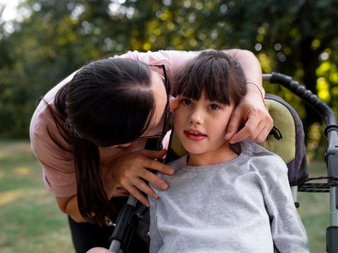 A woman gently kisses the cheek of a young girl sitting in a wheelchair outdoors, surrounded by greenery. The girl looks ahead with a soft expression while the woman shows affection.