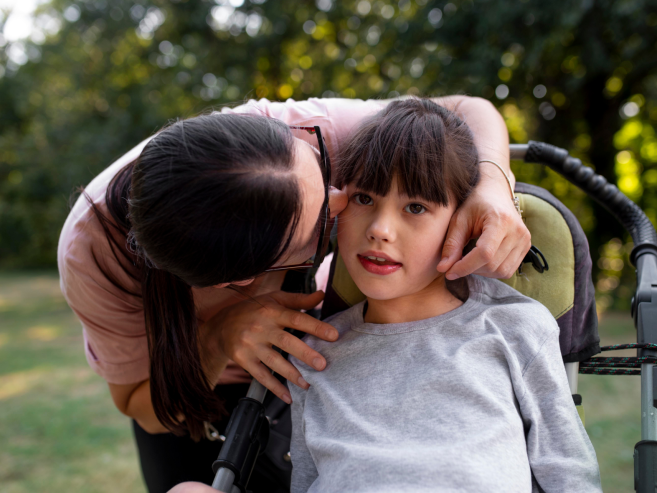 A woman gently kisses the cheek of a young girl sitting in a wheelchair outdoors, surrounded by greenery. The girl looks ahead with a soft expression while the woman shows affection.