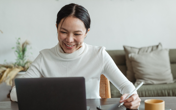 A woman sits at a table smiling at her laptop, holding a stylus and writing on a tablet. A yellow mug and some plants are visible in the background.