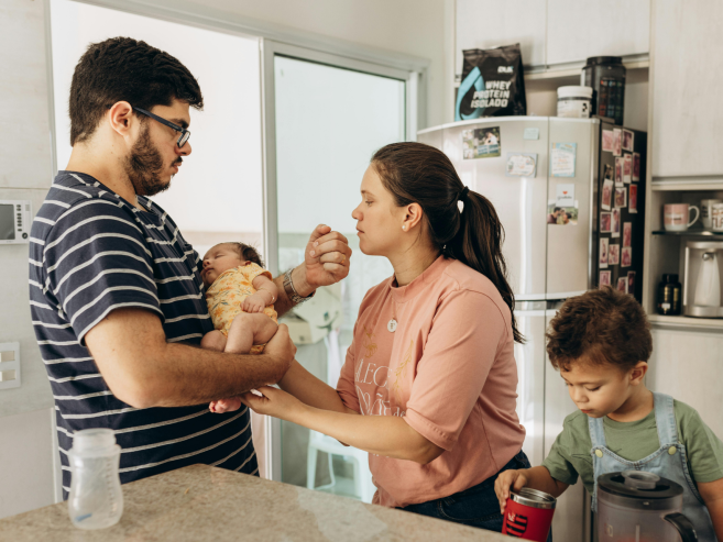 A man holds a sleeping baby while a woman stands close, touching the babys hand. A young child stands nearby at the counter, focused on a red container. They are all in a bright, modern kitchen.