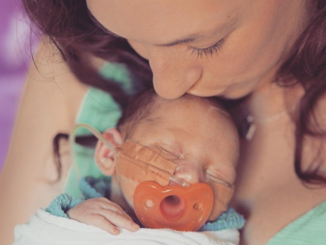 A woman gently kisses a newborn baby on the forehead. The baby, wrapped in a blanket, has a feeding tube and a heart-shaped pacifier. The background is softly blurred in purple tones.