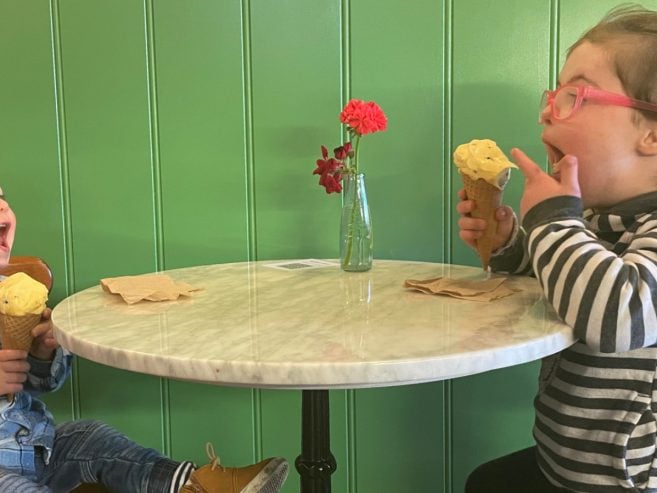 Two young children sit at a small round table with a green wall behind them, laughing and enjoying ice cream cones. A small vase with flowers sits on the table next to napkins.
