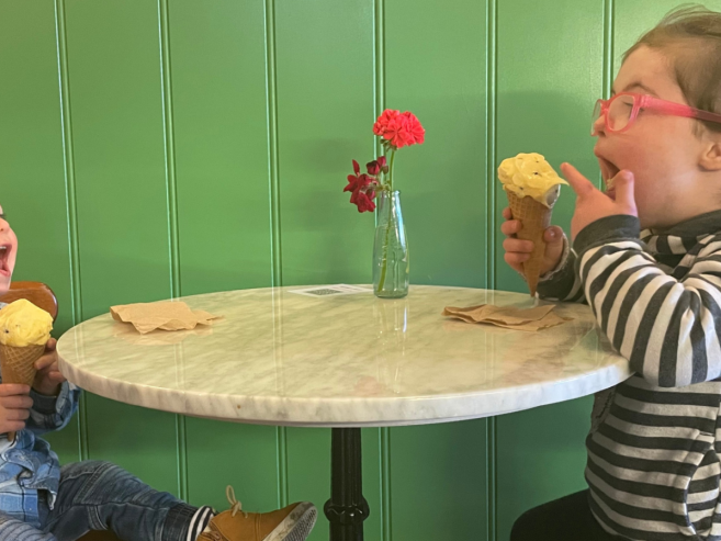 Two young children sit at a small round table with a green wall behind them, laughing and enjoying ice cream cones. A small vase with flowers sits on the table next to napkins.