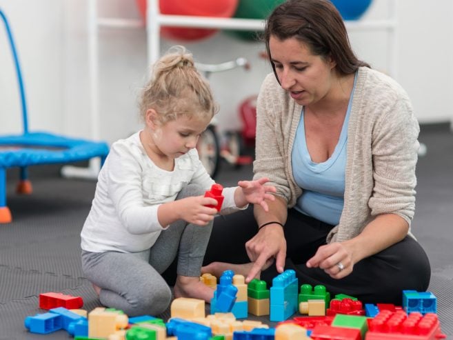 A woman and a young girl sit on the floor playing with colorful plastic building blocks. The girl is stacking blocks while the woman points and helps, both focused on the activity. Play equipment is visible in the background.