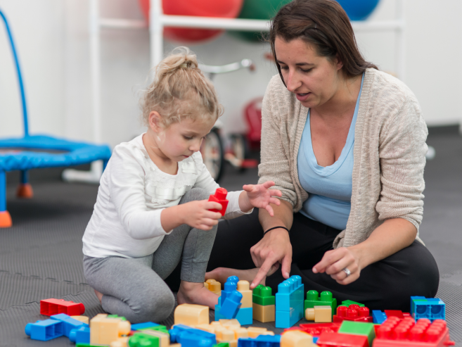A woman and a young girl sit on the floor playing with colorful plastic building blocks. The girl is stacking blocks while the woman points and helps, both focused on the activity. Play equipment is visible in the background.