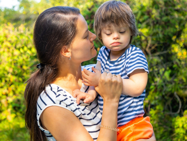 A woman holding a young child outdoors, both wearing striped shirts. The child looks down with a calm expression while the woman gazes at the child. Green foliage is visible in the background.
