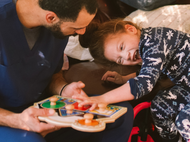 A man in scrubs helps a smiling child in a wheelchair play with a colorful wooden puzzle. The child wears star-patterned pajamas and leans happily towards the man. Soft sunlight fills the cozy room.