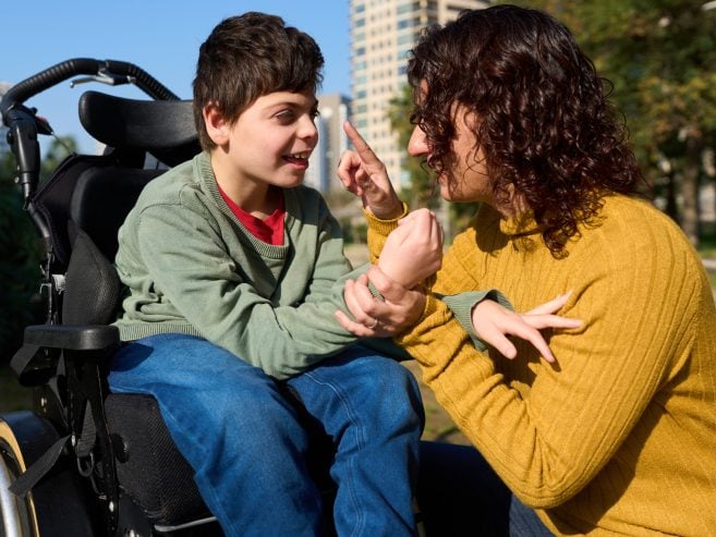 A woman in a yellow sweater communicates with a smiling boy in a wheelchair, using sign language outdoors on a sunny day, with buildings and trees in the background.