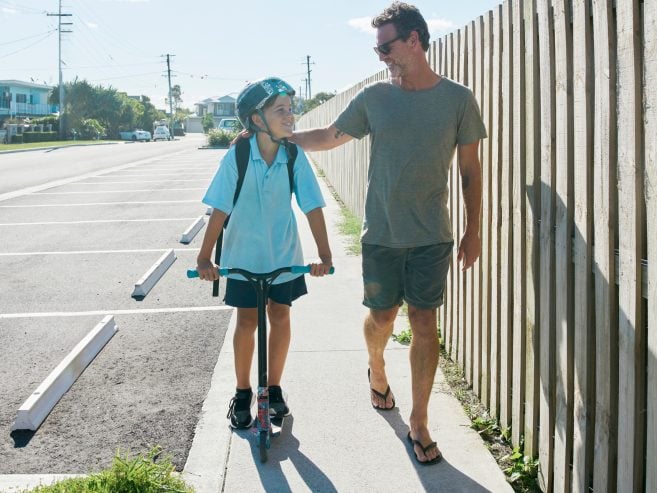 A smiling man walks beside a child riding a scooter on a sunny sidewalk. The child wears a helmet and backpack, while the man wears shorts, a t-shirt, and flip-flops, gently holding the childs shoulder.