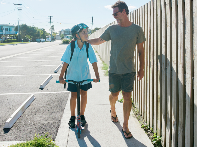 A smiling man walks beside a child riding a scooter on a sunny sidewalk. The child wears a helmet and backpack, while the man wears shorts, a t-shirt, and flip-flops, gently holding the childs shoulder.