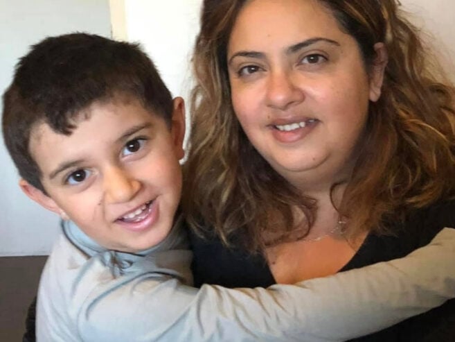 A young boy with short dark hair hugs a woman with long wavy hair and a black top. Both are smiling and looking at the camera, sitting closely together indoors.