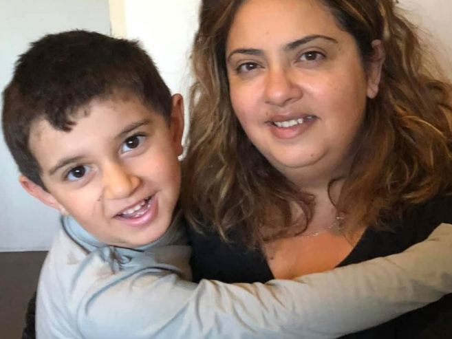 A young boy with short dark hair hugs a woman with long wavy hair and a black top. Both are smiling and looking at the camera, sitting closely together indoors.