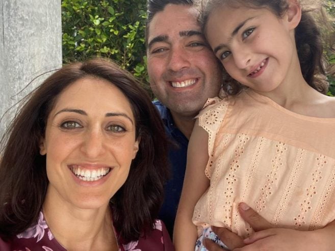 A woman, a man, and a young girl smile together for a selfie outdoors, with greenery in the background. The woman has shoulder-length brown hair, and the girl is wearing a peach-colored dress.