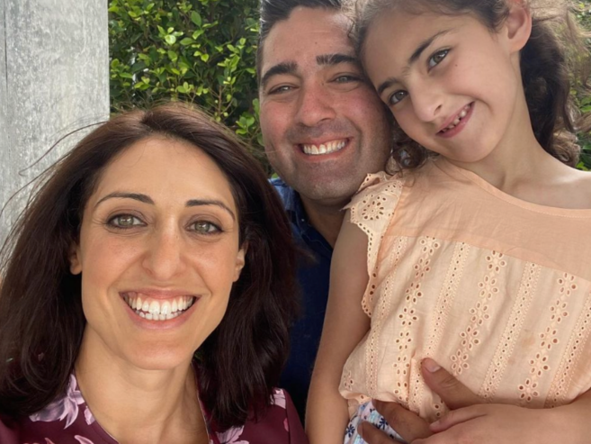 A woman, a man, and a young girl smile together for a selfie outdoors, with greenery in the background. The woman has shoulder-length brown hair, and the girl is wearing a peach-colored dress.