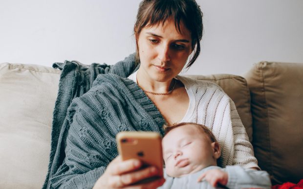 A woman sits on a couch with a gray blanket, holding a smartphone and looking at the screen while a sleeping baby rests on her lap, wrapped in a red blanket.