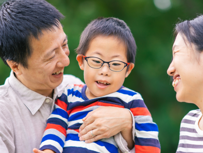 A smiling man and woman hold a young boy wearing glasses and a striped shirt, standing outdoors with a blurred green background. All appear happy and engaged with each other.