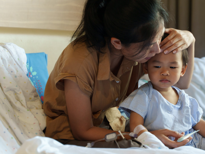 A woman gently kisses the forehead of a young child wearing hospital clothes, sitting together on a hospital bed with medical equipment and childrens books nearby.
