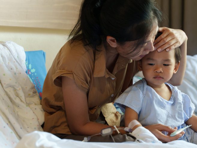 A woman gently kisses the forehead of a young child wearing hospital clothes, sitting together on a hospital bed with medical equipment and childrens books nearby.
