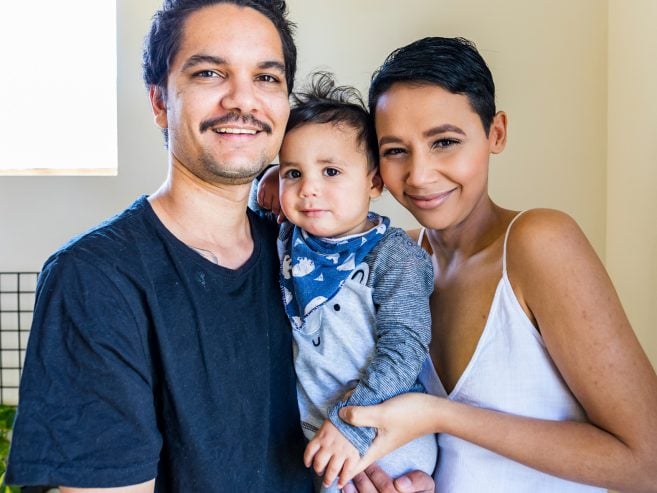 A smiling couple stands close together, holding their baby. The man wears a black shirt, the woman a white dress, and the baby is dressed in grey with a blue bib. They are indoors with natural light coming from a window.