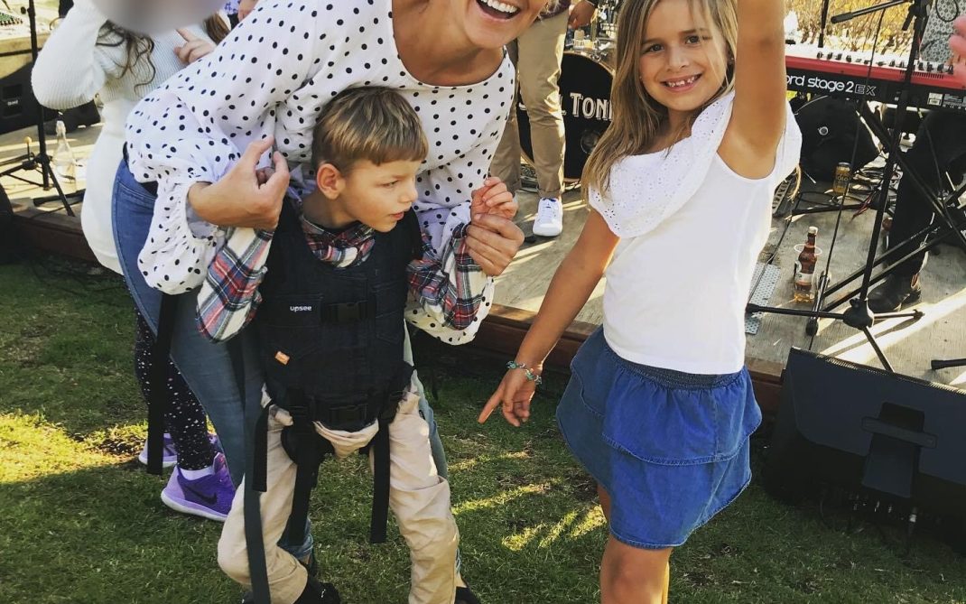 A smiling woman in sunglasses crouches beside a boy using a harness for support and a girl making a peace sign. Theyre outdoors at a live music event on grass, with instruments and blurred people in the background.