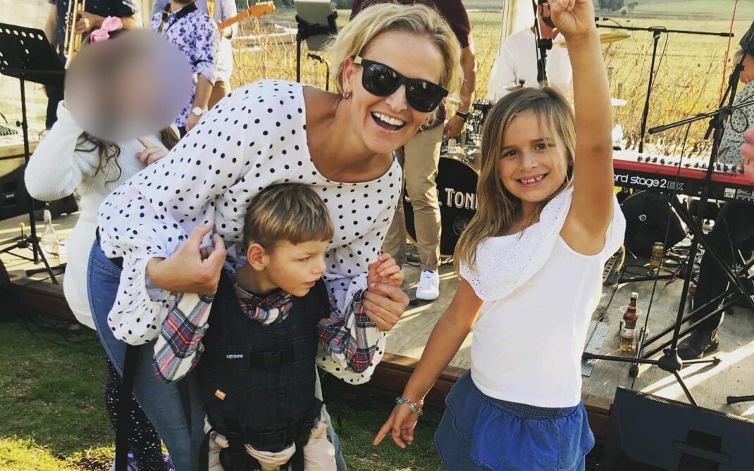 A smiling woman in sunglasses crouches beside a boy using a harness for support and a girl making a peace sign. Theyre outdoors at a live music event on grass, with instruments and blurred people in the background.