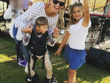 A smiling woman in sunglasses crouches beside a boy using a harness for support and a girl making a peace sign. Theyre outdoors at a live music event on grass, with instruments and blurred people in the background.
