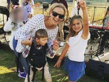 A smiling woman in sunglasses crouches beside a boy using a harness for support and a girl making a peace sign. Theyre outdoors at a live music event on grass, with instruments and blurred people in the background.