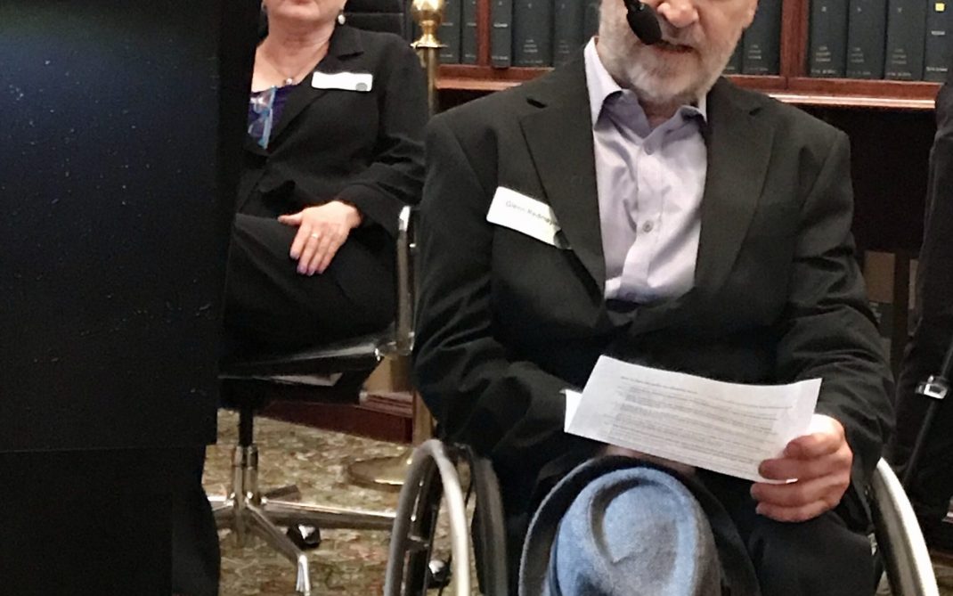 A man in a suit sits in a wheelchair, holding a sheet of paper and a hat, speaking into a microphone. He is in a library-like room with shelves of books in the background. A podium and another seated person are nearby.