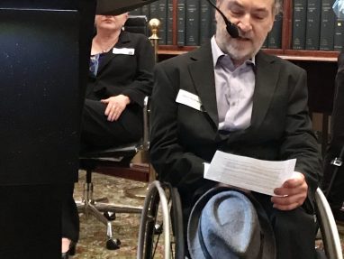 A man in a suit sits in a wheelchair, holding a sheet of paper and a hat, speaking into a microphone. He is in a library-like room with shelves of books in the background. A podium and another seated person are nearby.