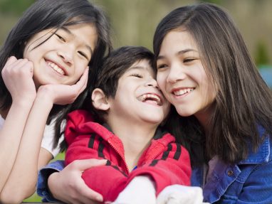 Three smiling children, two girls and one boy, are close together outdoors. The boy in the middle wears a red jacket, and all three appear happy and joyful, sharing a moment of laughter and affection.