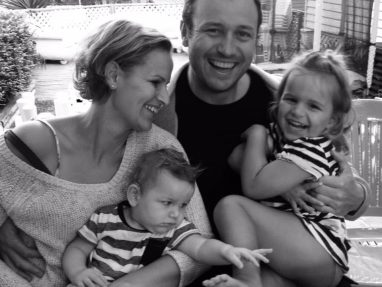 A smiling family of four sits closely together outdoors. The mother and father hold their two young children; the father and daughter smile at the camera, while the mother and son look at each other. The photo is in black and white.