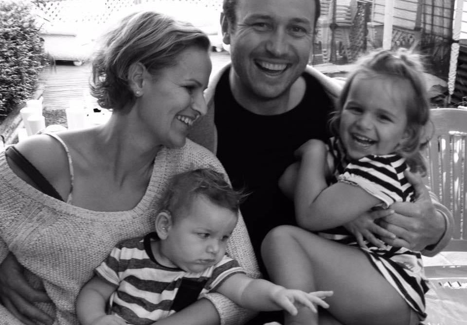 A smiling family of four sits closely together outdoors. The mother and father hold their two young children; the father and daughter smile at the camera, while the mother and son look at each other. The photo is in black and white.