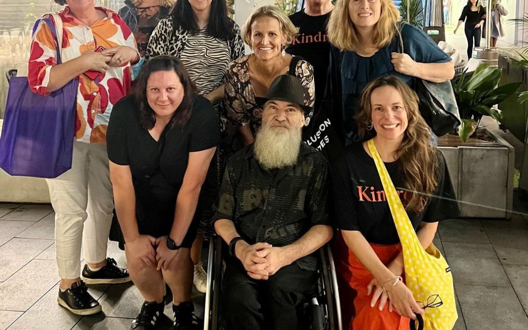 A group of nine people, including a man in a wheelchair, pose together and smile for the camera outdoors at night, surrounded by greenery and city lights.