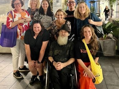 A group of nine people, including a man in a wheelchair, pose together and smile for the camera outdoors at night, surrounded by greenery and city lights.