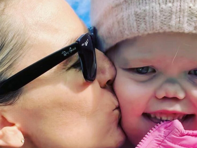 A woman wearing black sunglasses kisses a smiling young child dressed in a pink jacket and knit hat. The sky is blue in the background.