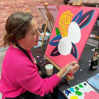 A woman in a bright pink sweater paints a colorful abstract floral design on a canvas at an art studio, with painting supplies and a bottle on the table beside her.