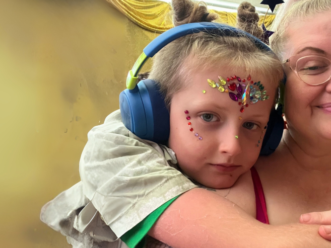 A child with headphones and face paint hugs an adult with glasses and a smile; both are close together in a warm, cozy setting with a yellow draped background.