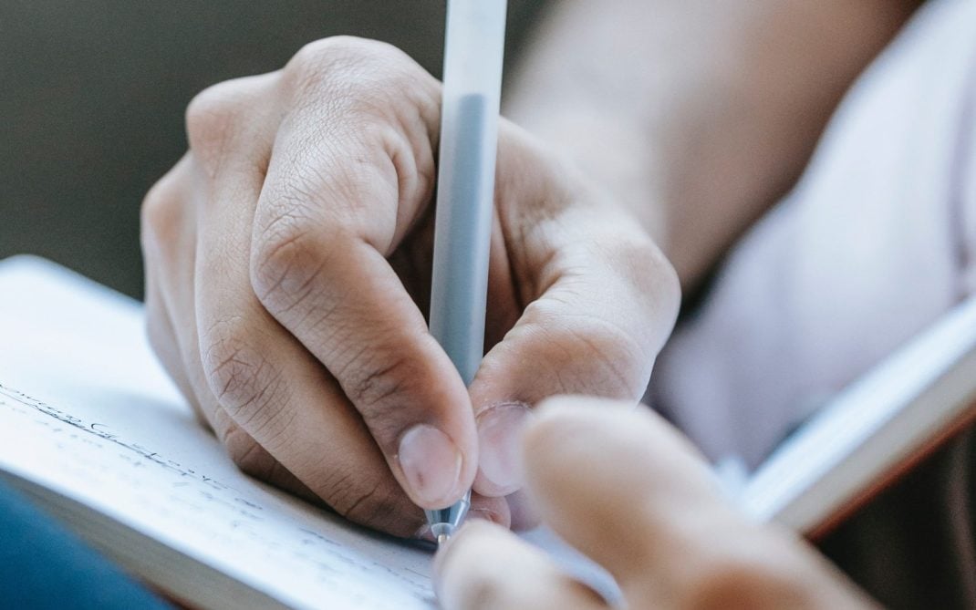 A close-up of a person’s hand writing in a notebook with a gray pen, with another hand holding the notebook. The background is softly blurred.