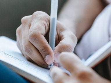 A close-up of a person’s hand writing in a notebook with a gray pen, with another hand holding the notebook. The background is softly blurred.