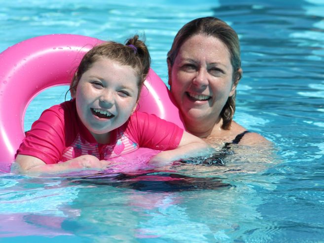 A smiling child with a pink swim ring and a woman stand in a clear blue swimming pool, both enjoying the water on a sunny day.