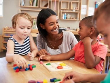 A teacher smiles while sitting at a table with three young children playing with colorful wooden blocks in a classroom setting.