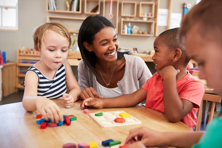 A teacher smiles while sitting at a table with three young children playing with colorful wooden blocks in a classroom setting.