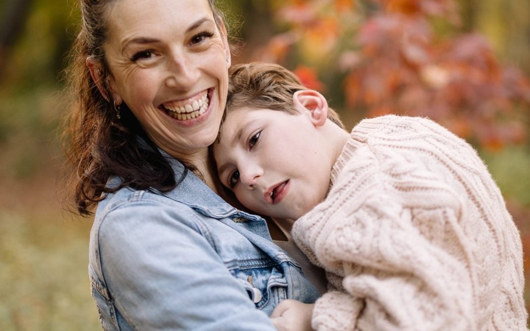 A smiling woman in a denim jacket holds a young boy wearing a cream sweater in her arms. They are outdoors in a forest with green and autumn-colored leaves in the background.