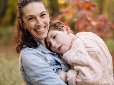 A smiling woman in a denim jacket holds a young boy wearing a cream sweater in her arms. They are outdoors in a forest with green and autumn-colored leaves in the background.